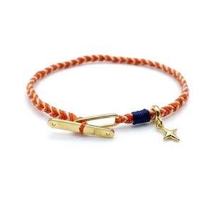 Op de afbeelding: Een gevlochten armband van oranje en wit koord, vastgezet met een goudkleurige metalen knevelsluiting. Een kleine donkerblauwe draadwikkeling zit bij de sluiting en een goudkleurige vierpuntige sterbedel hangt aan een kleine ring.
