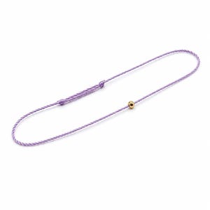Könnte beinhalten: Ein zartes Armband aus lavendelfarbenem Kordelmaterial mit einer einzelnen polierten goldfarbenen Perle und einem verstellbaren Schiebeknotenverschluss. Der dünne, gedrehte fliederfarbene Faden bildet eine durchgehende Schlaufe, akzentuiert durch die kleine, runde goldene Metallperle.