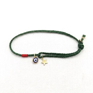Puede incluir: Una pulsera de cordón trenzado verde oscuro con un pequeño dije de ojo turco dorado con esmalte azul y blanco, un dije de estrella dorado y un acento de hilo rojo cerca del ojo, asegurada con un cierre de nudo.