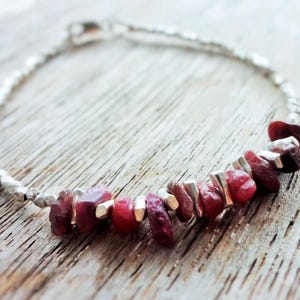 Könnte beinhalten: Ein silbernes Armband mit einer Kette aus kleinen, facettierten Perlen und einer Reihe unregelmäßiger, tiefroter Edelsteine. Das Armband liegt auf einer Holzoberfläche und hebt die natürlichen Farbtöne des Schmucks hervor.