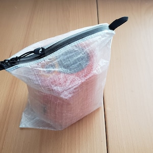 Könnte beinhalten: Ein transparenter Beutel mit schwarzem Reißverschluss und Zuglasche. Der Beutel enthält ein rotes Objekt, möglicherweise eine Rolle Klebeband oder Stoff. Der Beutel liegt auf einer hellbraunen Holzoberfläche.