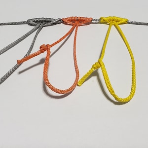 Puede incluir: Tres bucles de cuerda en gris, naranja y amarillo están conectados a una cuerda gris. Cada bucle está atado con un nudo. El fondo es blanco.