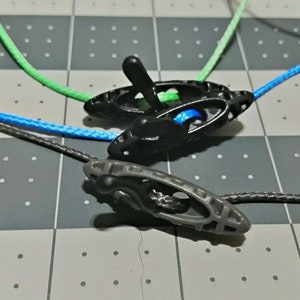 May include: Close-up of three cord locks with colorful cords. Two black cord locks and one gray cord lock are visible. The cords are blue, green, and gray. The cord locks are made of plastic and have a spring-loaded mechanism.