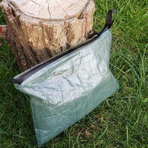 May include: A light green pouch with a black zip and a black loop. The pouch is made of a crinkled, translucent material. The pouch is on green grass next to a tree stump. The pouch is approximately 25 cm long.