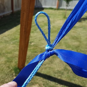 May include: Close-up of a blue rope and webbing knot, likely for a hammock or swing. The knot is tied with a loop and is connected to a blue strap. The background includes a wooden post and green grass.