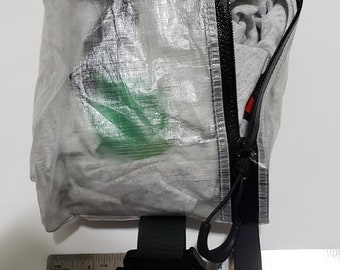 Puede incluir: Una bolsa transparente y rectangular con detalles negros, que contiene artículos grises. La bolsa tiene una cremallera negra y una correa negra. Una regla con pulgadas está al lado de la bolsa.