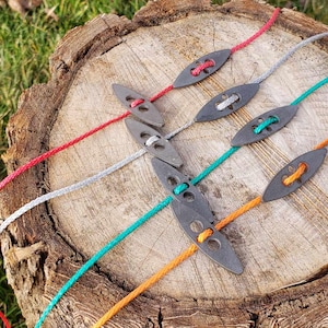 May include: A collection of slingshot accessories, featuring gray leather pouches with colorful cords in red, white, green, and orange. The accessories are arranged on a weathered wooden stump, set against a backdrop of green grass.