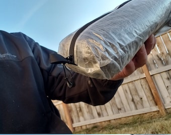 Op de afbeelding: Een opgerolde, doorschijnende, grijze en zwarte ultralichte stuff sack met een zwarte ritssluiting en trekkoord. De stuff sack wordt voor een houten hek en blauwe lucht gehouden.