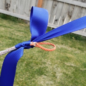 May include: Close-up of a blue strap and white rope knotted together. An orange loop is attached to the knot. The background is a green lawn and a wooden fence.