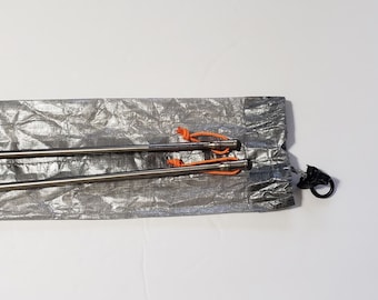 Op de afbeelding: Twee roestvrijstalen tentpinnen met oranje koorden, opgeborgen in een zilveren, gekreukelde zak. De pinnen zijn lang en puntig, ontworpen om tenten of andere outdooruitrusting vast te zetten. Een zwarte clip is aan de zak bevestigd.