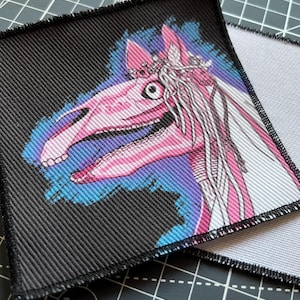 May include: Two square fabric patches. One patch features a pink and white horse head with a crown, set against a black background with a blue and purple outline. The second patch is partially visible.