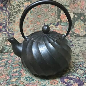 May include: A black cast iron teapot with a spiral design. The teapot has a curved handle and a small knob on the lid. The spout is curved and angled. The teapot is a traditional style and is likely used for brewing tea.