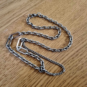 May include: A silver chain necklace with two different chain styles. The necklace has a unique clasp design with two interlocking pieces.
