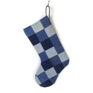 May include: A Christmas stocking with a checkered pattern in shades of blue denim. The stocking is hanging from a silver hook against a white background. The checkered pattern is made up of squares of varying shades of blue.