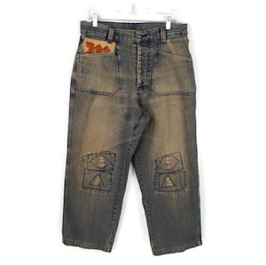 May include: Vintage blue denim jeans with a faded wash. Features include front and back pockets, decorative patches on the knees, and a unique pocket detail. The jeans are hung on a white hanger.