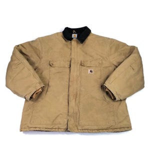 May include: A tan work jacket with a black corduroy collar. The jacket has two chest pockets and two lower pockets. The Carhartt logo is visible on the right chest pocket. The jacket is made of a durable, heavy-duty material.