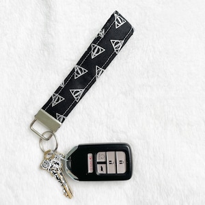 May include: Black fabric wristlet key fob with white Deathly Hallows symbols. Attached to a silver key ring with a car key and a decorative key. The key fob is approximately 1 inch wide and 6 inches long.
