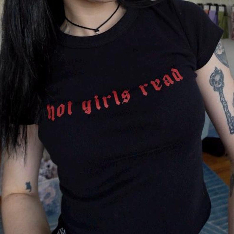 以下が含まれることがあります： 赤いゴシック体の「hot girls read」という文字が入った黒いTシャツ。Tシャツはクルーネックで半袖です。画像はTシャツを着ている人の上半身を示しています。