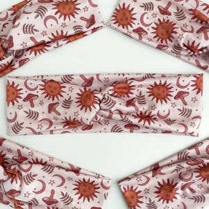 May include: Light pink headbands with a repeating pattern of suns, moons, mushrooms, and stars in shades of orange and brown. The headbands are knotted at the center. The image shows several headbands.