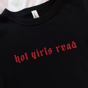 以下が含まれることがあります： 赤いゴシック体の「hot girls read」というフレーズがプリントされた黒いTシャツ。シャツはクルーネックと半袖です。タグには「Canvas」と書かれています。