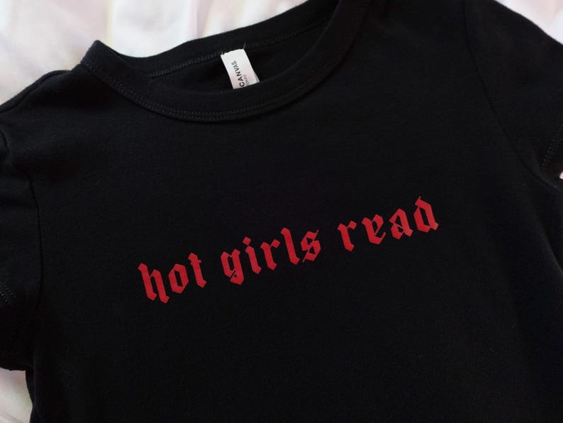 以下が含まれることがあります： 赤いゴシック体の「hot girls read」というフレーズがプリントされた黒いTシャツ。シャツはクルーネックと半袖です。タグには「Canvas」と書かれています。