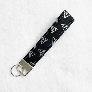 May include: Black fabric wristlet key fob with a silver metal key ring. The key fob features a repeating white graphic pattern of a triangle with a vertical line through it. The key fob is approximately 6 inches long.