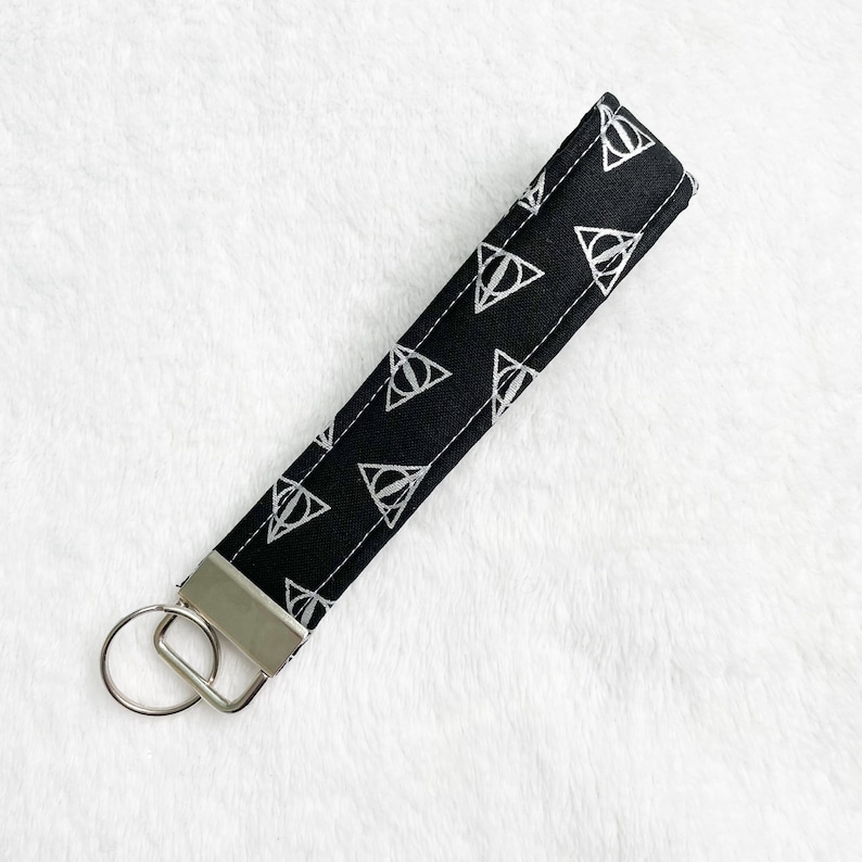 May include: Black fabric wristlet key fob with a silver metal key ring. The key fob features a repeating white graphic pattern of a triangle with a vertical line through it. The key fob is approximately 6 inches long.