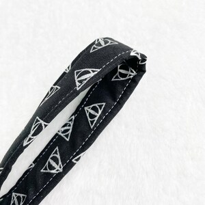 May include: Black fabric strap with a repeating white Deathly Hallows symbol pattern. The strap is stitched with white thread along the edges. The background is a soft, white, textured surface.