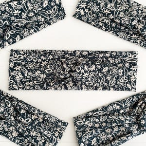 May include: Several dark navy headbands with a cream floral pattern are arranged on a white surface. The headbands are twisted at the top. The floral design includes leaves and flowers. The headbands are a stylish accessory.