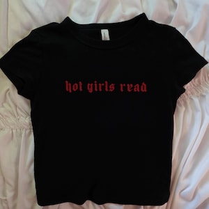 以下が含まれることがあります： 胸に赤いゴシック体の文字で「hot girls read」と書かれた黒い半袖Tシャツ。 シャツはクルーネックで、柔らかい素材でできています。 シャツは白い表面に置かれています。