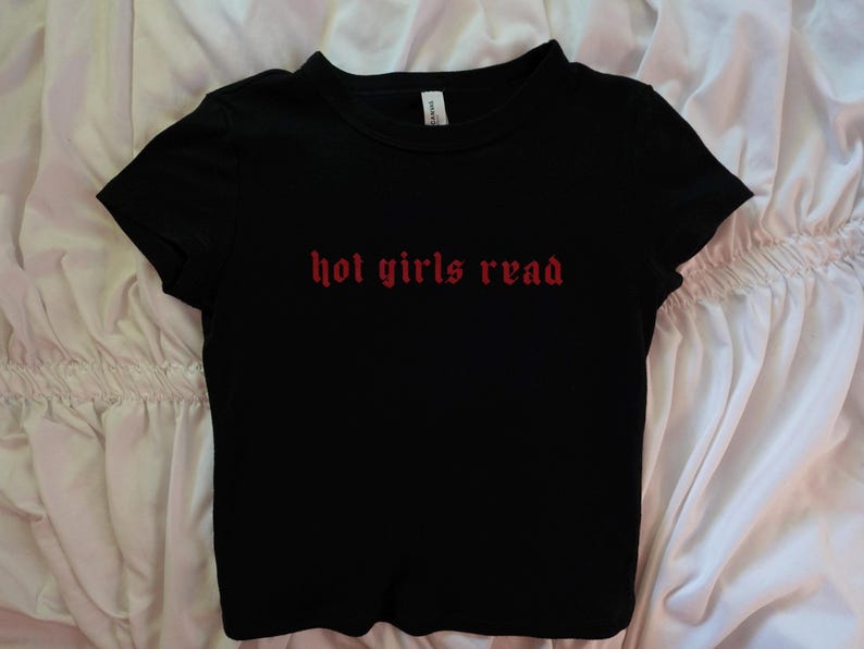 以下が含まれることがあります： 胸に赤いゴシック体の文字で「hot girls read」と書かれた黒い半袖Tシャツ。 シャツはクルーネックで、柔らかい素材でできています。 シャツは白い表面に置かれています。