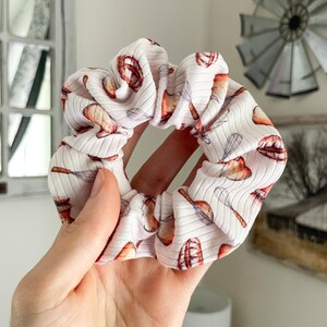 May include: A white ribbed fabric hair scrunchie featuring a repeating pattern of brown and orange kitchen whisks and mixing bowls. The accessory has a gathered, elasticized design with a soft texture and a consistent print throughout the material.