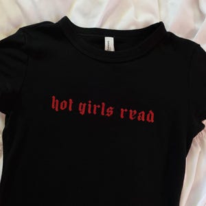 以下が含まれることがあります： 赤い文字で「hot girls read」と書かれた黒いTシャツ。Tシャツは半袖で、クルーネックです。テキストはゴシック体です。Tシャツは柔らかい素材で作られています。