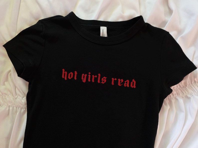 以下が含まれることがあります： 赤い文字で「hot girls read」と書かれた黒いTシャツ。Tシャツは半袖で、クルーネックです。テキストはゴシック体です。Tシャツは柔らかい素材で作られています。