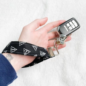 May include: Black wrist lanyard with white triangle symbols. The lanyard is attached to a car key fob with a silver metal clasp. The background is a soft, white, textured surface.