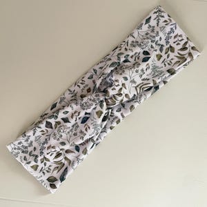 May include: White headband with a botanical print featuring green and gray leaves and sprigs. The fabric is knotted at the center. This accessory is suitable for various hairstyles and adds a touch of nature-inspired style.
