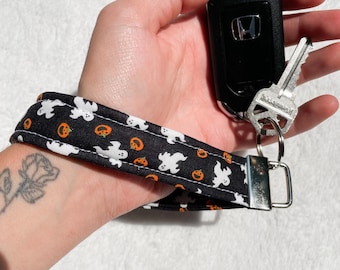 May include: Black Halloween-themed wristlet key fob with white ghost and orange pumpkin patterns. The key fob is attached to a silver metal ring holding a car key and a car key fob.