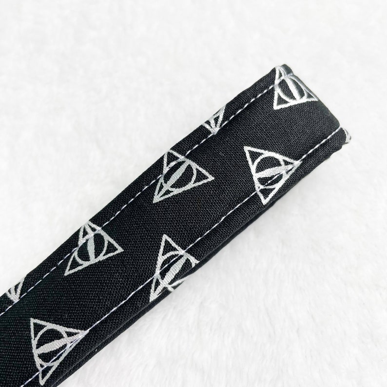 May include: Black fabric strap with a repeating silver Deathly Hallows symbol pattern. The strap is stitched with white thread along the edges. The background is a soft, white, textured surface.