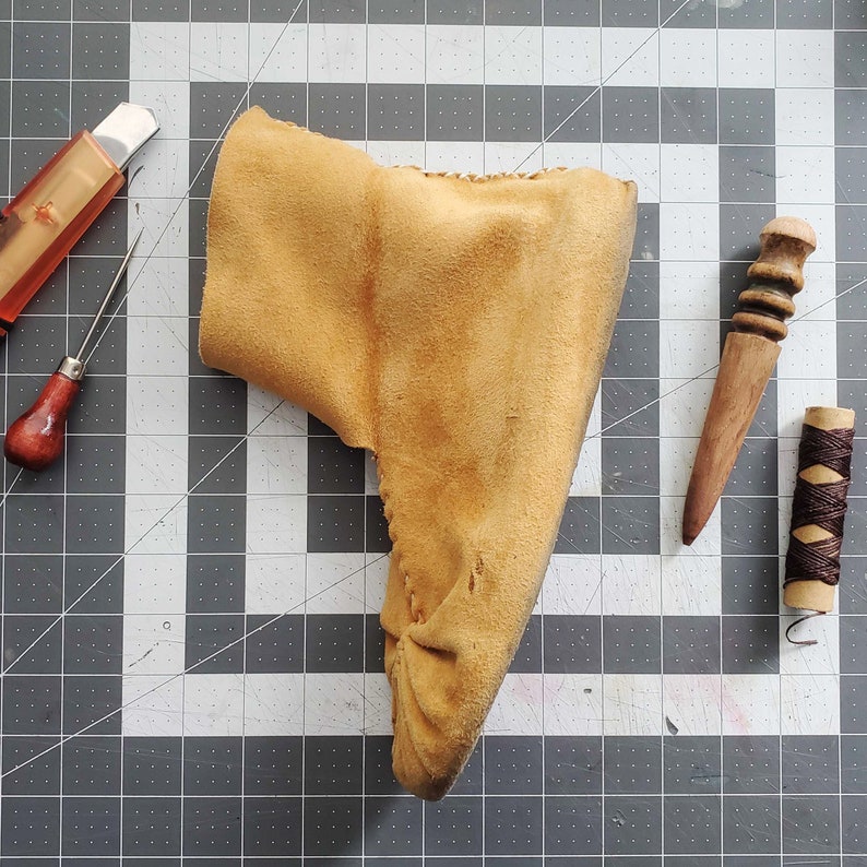 May include: A close-up of a tan leather moccasin with a pointed toe. The moccasin is on a gray and white checkered surface.