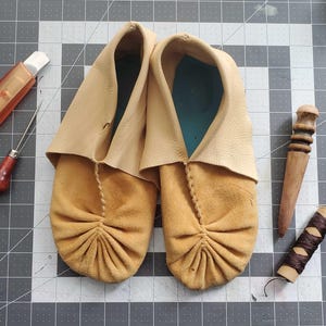 May include: A pair of tan leather moccasins with a folded cuff. The moccasins are made of soft, supple leather and have a simple, elegant design.