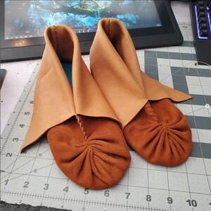 May include: A pair of brown leather moccasins with folded-over tops. The moccasins have a suede sole and are made with a decorative stitch.