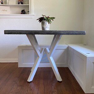 May include: A gray concrete table with a white X-shaped base. The table is set against a white wall and has a small potted plant on top.