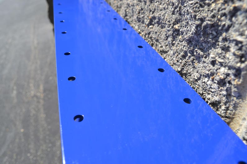 May include: A close-up of a blue metal sheet with evenly spaced holes drilled along the length. The sheet is attached to a rough, gray concrete wall.