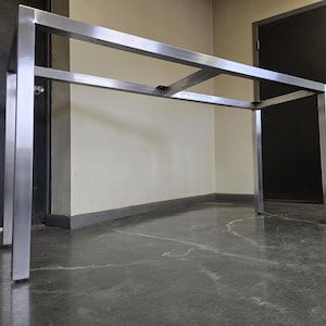 May include: A silver metal table base with a rectangular top and four legs. The table base is standing on a gray concrete floor.