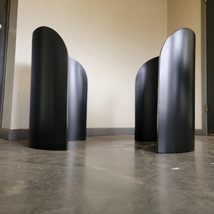 May include: Two black metal table bases with a curved design. The bases are standing on a concrete floor.