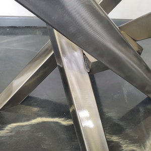 May include: Close-up of a metal table base with four legs that intersect in the center. The legs are made of polished stainless steel and have a smooth, reflective surface.