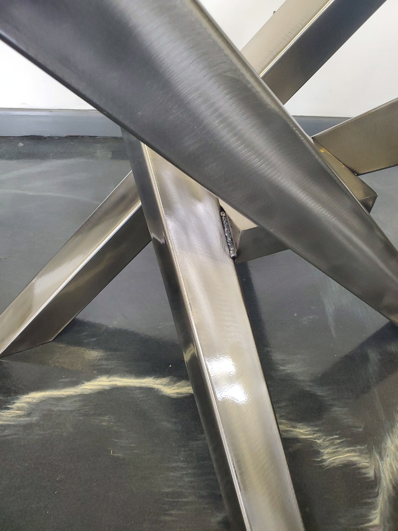 May include: Close-up of a metal table base with four legs that intersect in the center. The legs are made of polished stainless steel and have a smooth, reflective surface.