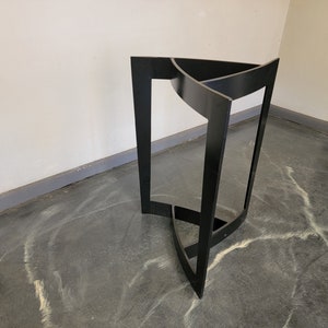 May include: Black metal table base with a triangular design. The base is made of three pieces of metal that are joined together to form a triangle. The base is standing on a grey concrete floor.