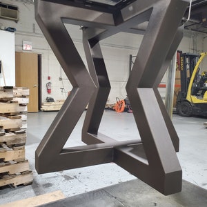 May include: A dark brown metal table base with a geometric design. The base is suspended in mid-air in a warehouse setting.