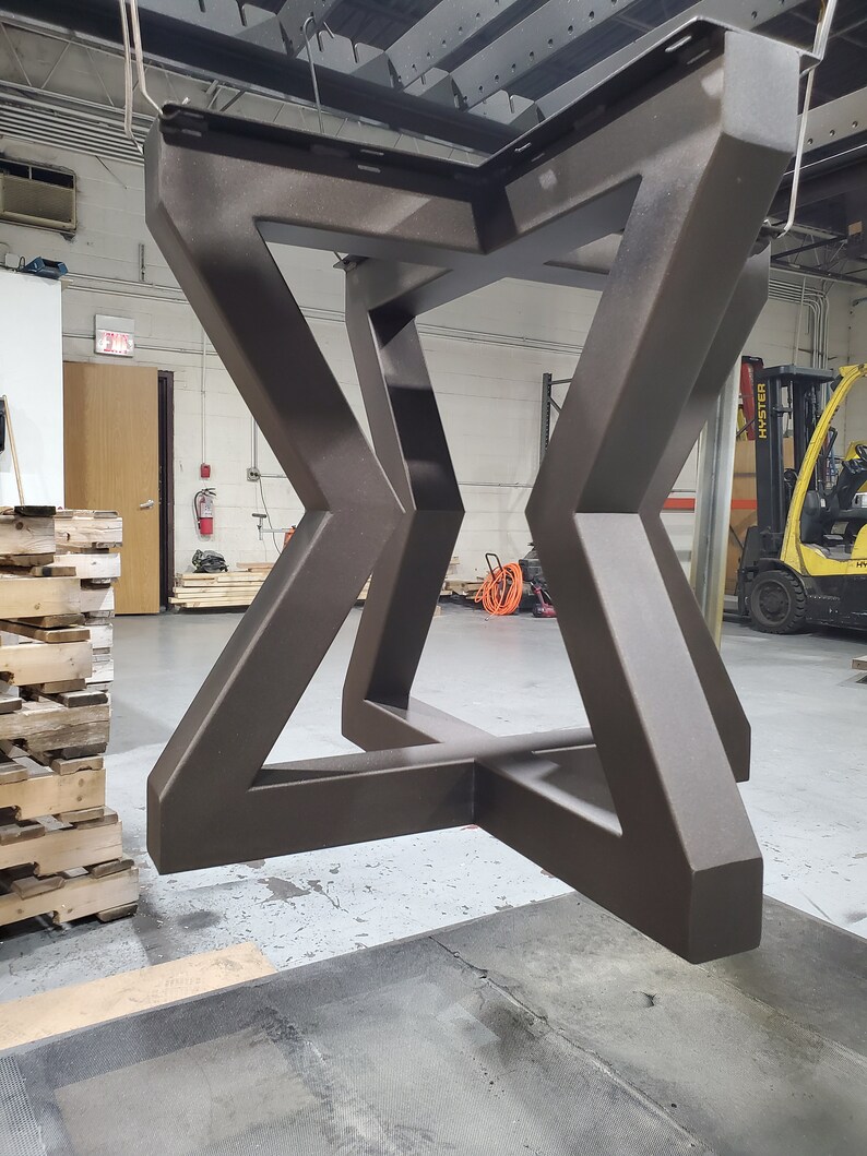 May include: A dark brown metal table base with a geometric design. The base is suspended in mid-air in a warehouse setting.